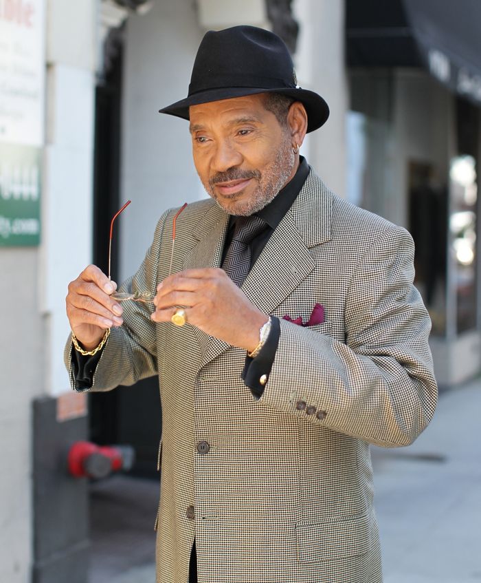 An elegant senior man in a formal coat and fedora smiling outdoors, adjusting his eyeglasses.