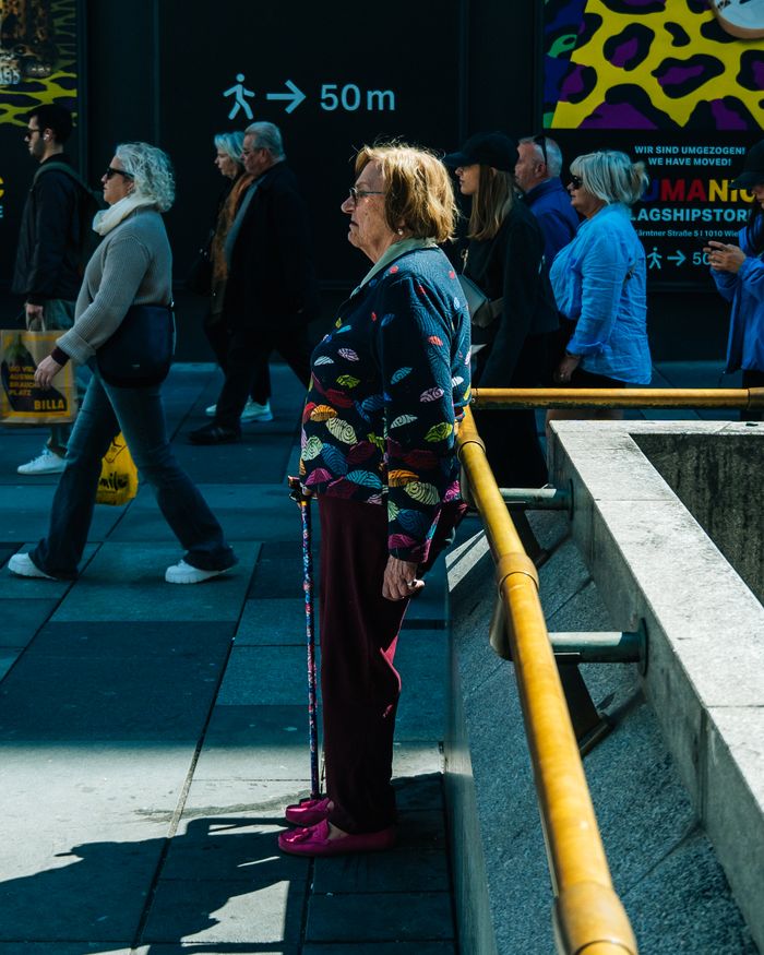 Senior woman stands with a cane on a bustling urban sidewalk, contrasting the crowd.