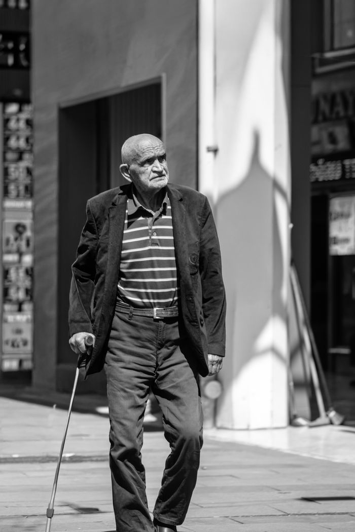 Black and white photo of an elderly man walking with a cane in a city environment during the day.