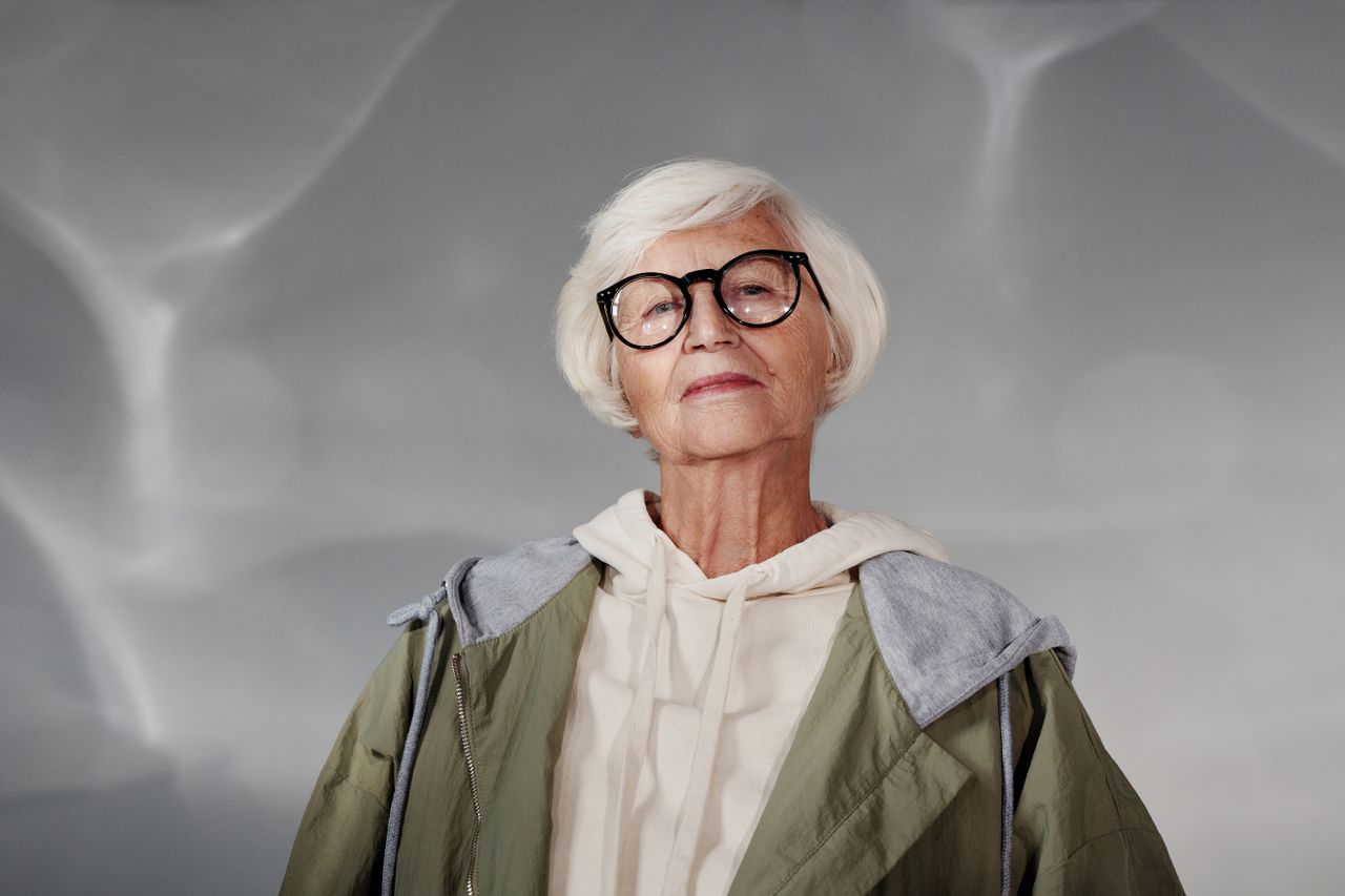 Portrait of a confident senior woman with white hair, wearing eyeglasses and a stylish jacket, posing indoors.