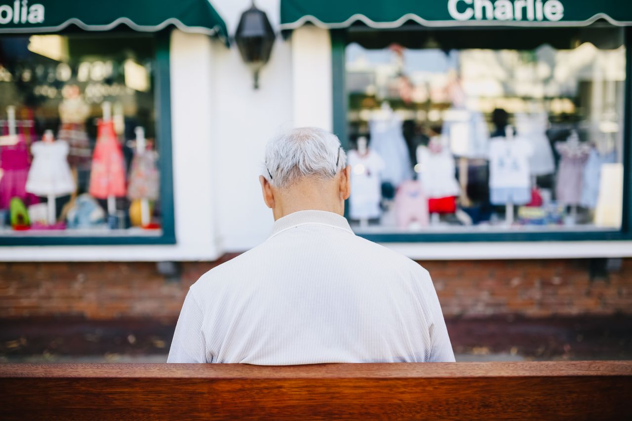 Senior man with white hair sitting on a bench facing a clothing store in an urban setting.