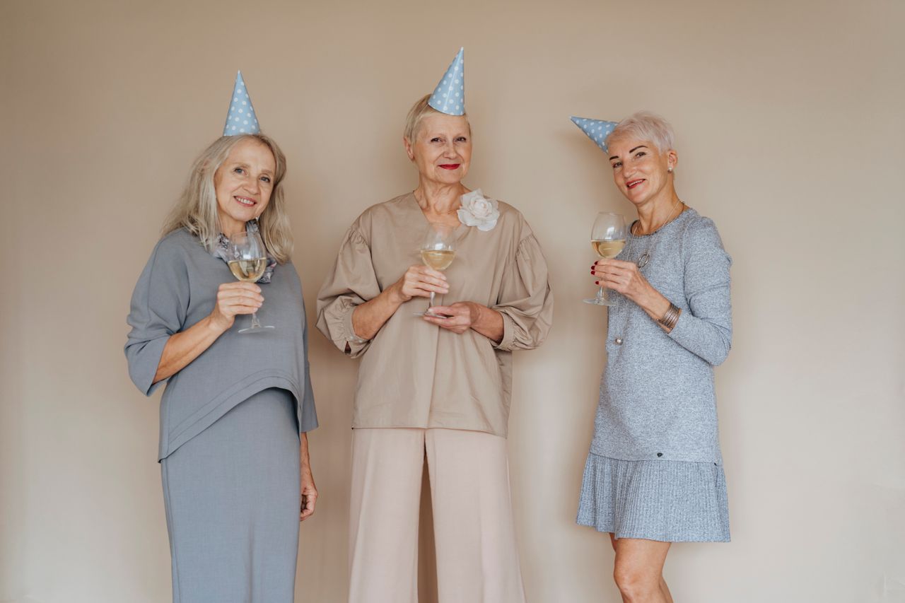 about-me-03 Three joyful senior women in party hats with glasses of wine celebrating indoors.