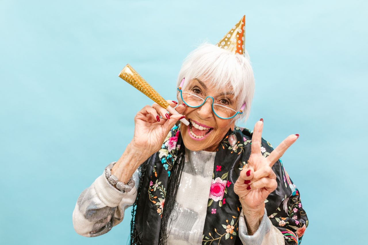 Senior woman in party hat joyfully celebrating, making peace sign on a light blue background.
